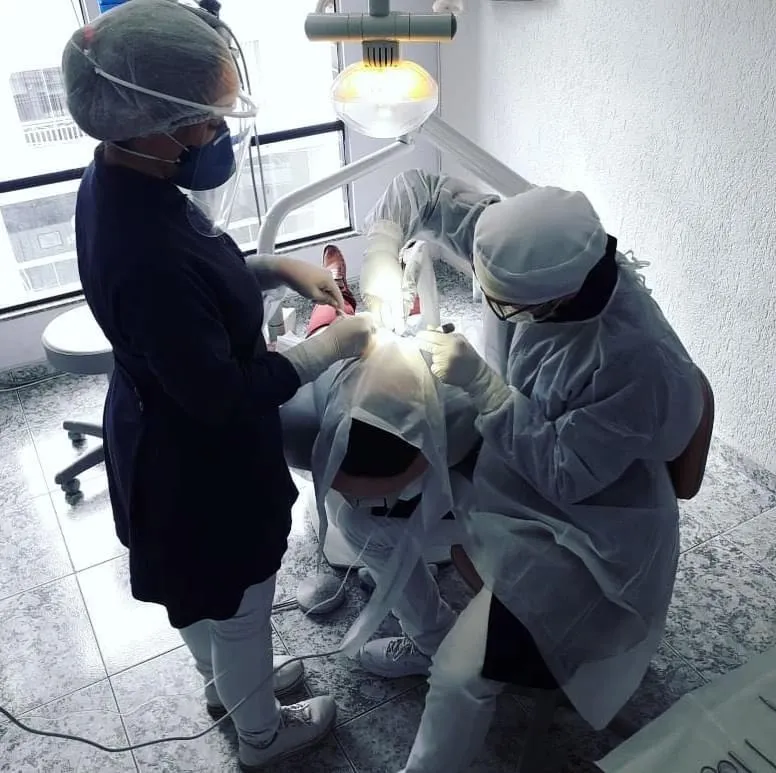 Atendimento no consultório da Patrocínio Odontologia: duas profissionais com máscara, touca e avental atendem um paciente na cadeira odontológica.