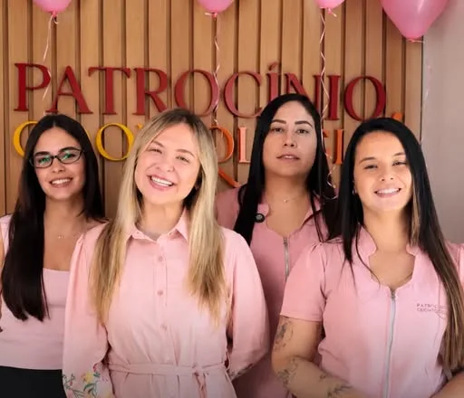 Quatro integrantes da equipe da Patrocínio Odontologia, de uniforme rosa, sorrindo em frente ao letreiro da clínica decorado com balões.